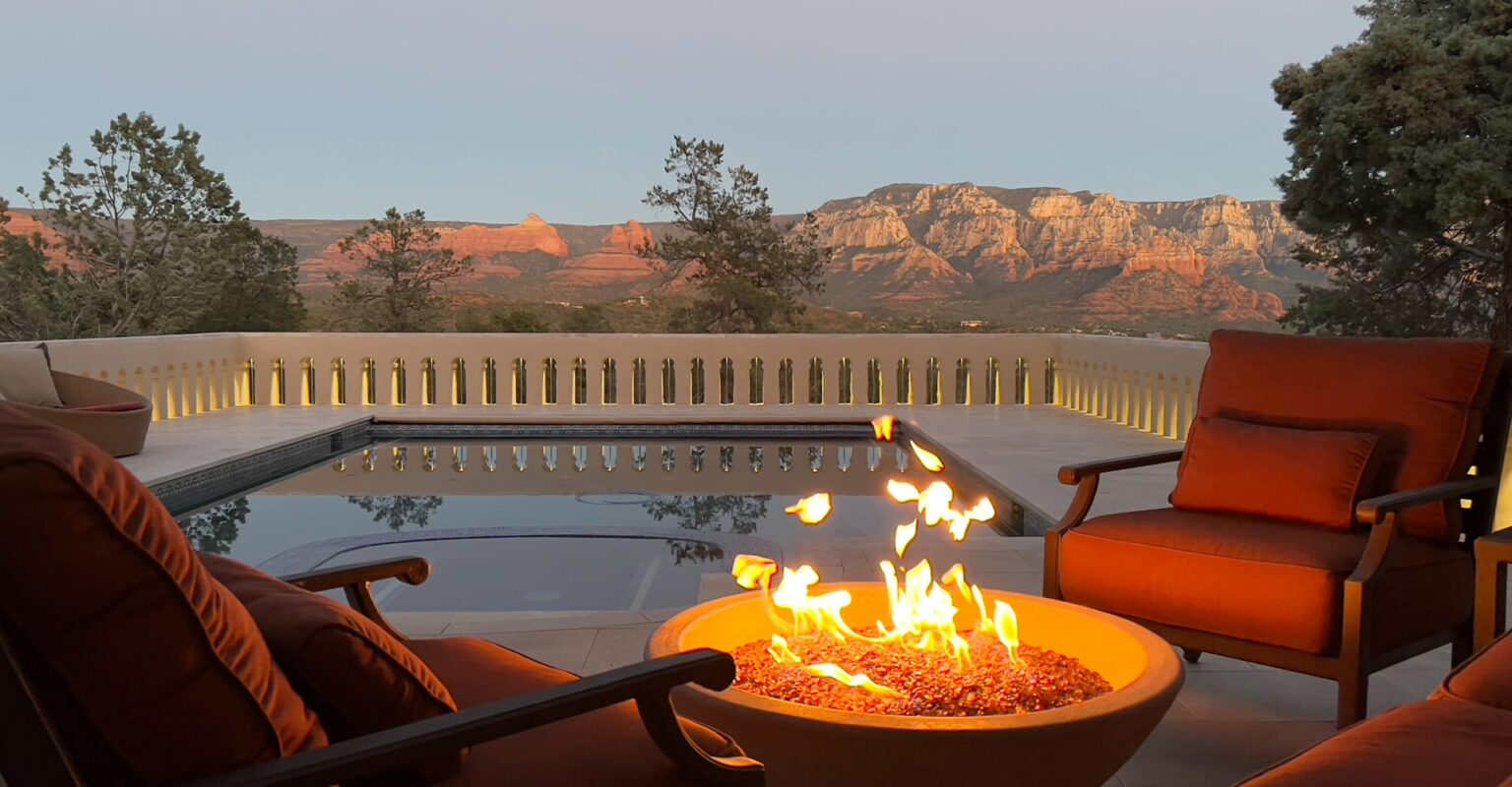 Remodeled Sedona backyard with a swimming pool, fire pit seating, illuminated stucco railing, mature trees, and panoramic red rock views at sunset.